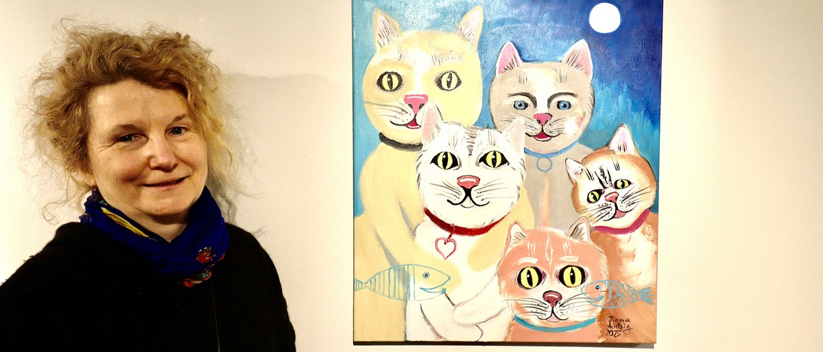 Die Künstlerin steht vor ihrem Gemälde. Auf dem Gemälde sind Katzen zu sehen.