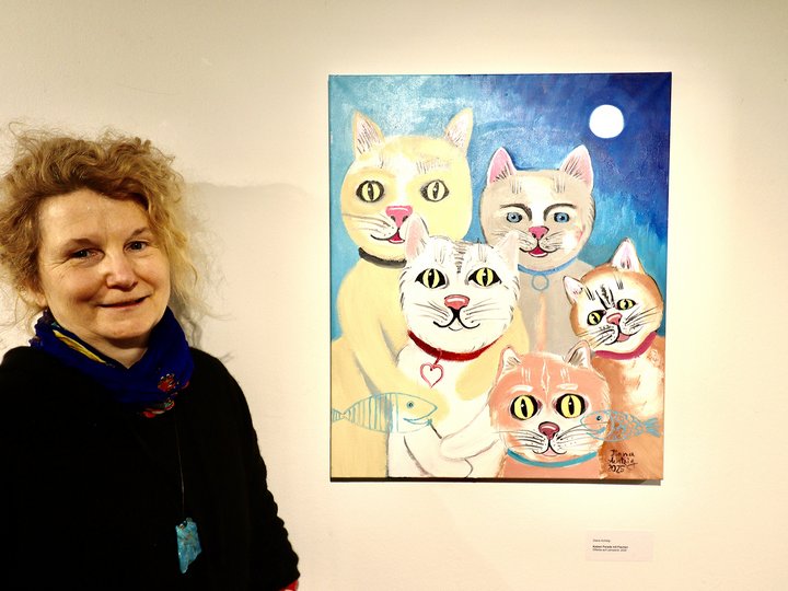Die Künstlerin steht vor ihrem Gemälde. Auf dem Gemälde sind Katzen zu sehen.